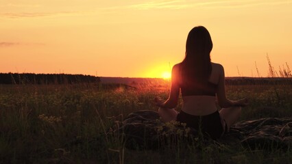 Young yogi woman sits on grass and meditates in lotus position against sunset. Dawn morning. Sports girl relaxes after workout outdoors in park. Yoga asana for meditation. Nature. Healthy lifestyle