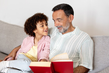 Senior Latin grandfather reading book to his grandchild boy indoor