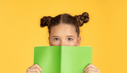 Curious young girl with buns hairstyle peeks over a green book, her eyes wide with intrigue