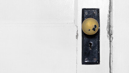 Horizontal color photo of a dirty tarnished and damaged gold doorknob on a white wooden interior door with cracked and peeling paint. Copy space