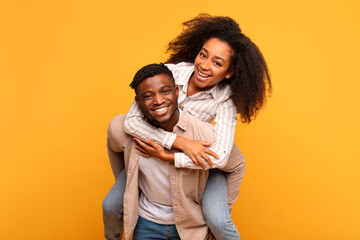 Happy black couple piggybacking with joy on yellow background