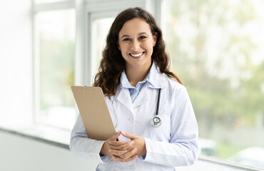 Cheerful lady doctor in white coat, with stethoscope and clipboard - obrazy, fototapety, plakaty