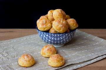 Ceramic bowl filled with choux pastry balls with cheese, wooden table, black background. French food, gougères.