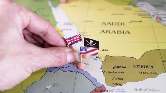 Hand placing pins adorned with the flags of the USA, UK, European union and Israle surrounding a pirate insignia onto a map of the Red Sea region. It symbolically represents the intricate geopolitical