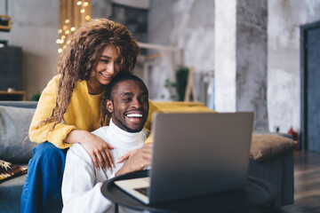 Joyful African-American couple home interior at winter, woman affectionately hugging man from behind while engage with laptop computer. Symbolizing love, comfort, and digital lifestyle