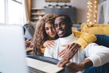 Young adult interracial couple enjoying leisure time at home, with a Black man and a Middle-Eastern woman smiling and browsing internet together on laptop, depicting love, technology, cozy lifestyle