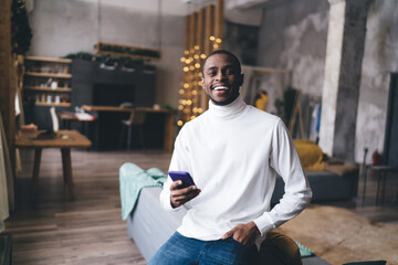 Portrait of a smiling Black man holding a smartphone in a stylish living room, exemplifying a relaxed, contemporary lifestyle and the integration of technology into daily life