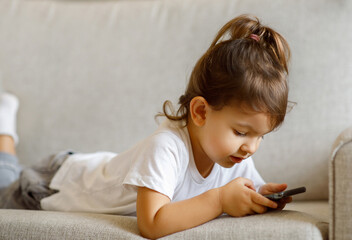 Fototapeta premium Cute Little Girl Playing Online Games On Smartphone While Lying On Couch