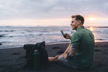 Smiling male with smartphone in hand on sandy seashore