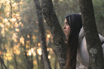 Un momento de paz y conexión con la naturaleza: una joven disfruta de la belleza del bosque al atardecer, apoyada en un árbol que le ofrece sombra y consuelo - obrazy, fototapety, plakaty