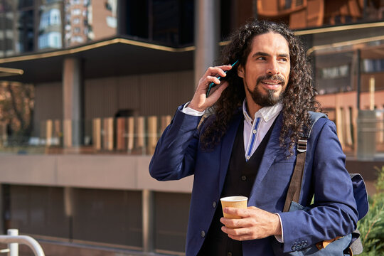 Smiling mature Latin businessman with long hair on a phone call, holding a coffee cup outdoors in the city
