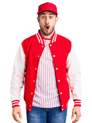 Young handsome man wearing baseball jacket and cap scared and amazed with open mouth for surprise, disbelief face