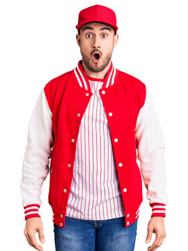 Young handsome man wearing baseball jacket and cap scared and amazed with open mouth for surprise, disbelief face