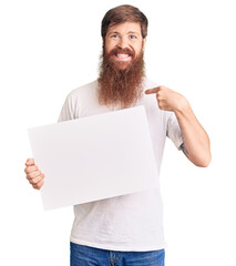 Handsome young red head man with long beard holding blank empty banner pointing finger to one self smiling happy and proud