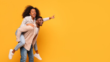 Joyful black couple in casual clothes enjoying piggyback ride on yellow background