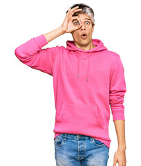 Fototapeta premium Young hispanic man wearing casual pink sweatshirt doing ok gesture shocked with surprised face, eye looking through fingers. unbelieving expression.