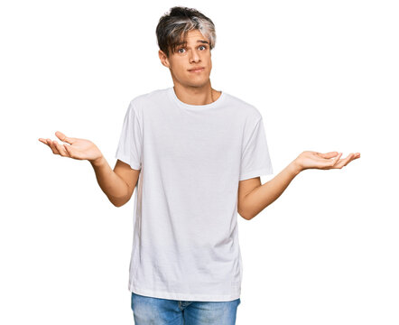 Young hispanic man wearing casual white tshirt clueless and confused expression with arms and hands raised. doubt concept.