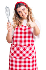 Young caucasian woman wearing apron holding whisk smiling happy and positive, thumb up doing excellent and approval sign