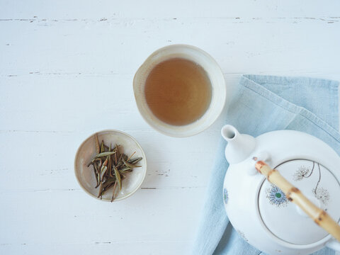 Tea cup Chinese white tea called silver needle or Bai Hao Yin Zhen. Leaf buds infused in bowl and hot beverage served in porcelain on white wooden table next to teapot on blue cloth. Top view.