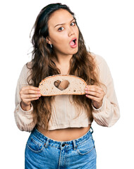 Young hispanic girl holding bread with heart symbol in shock face, looking skeptical and sarcastic,...