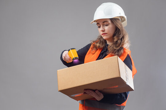 Woman warehouse worker. Storekeeper girl with DCT. Woman scans barcode on box. Storekeeper with parcel in hands. Girl registers parcel in database. Storekeeper on gray. Warehouse worker in hardhat