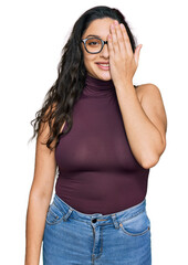 Brunette young woman wearing casual clothes and glasses covering one eye with hand, confident smile on face and surprise emotion.