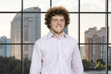 Happy young attractive man with curly hair bites his lip. Indoor window in the background.