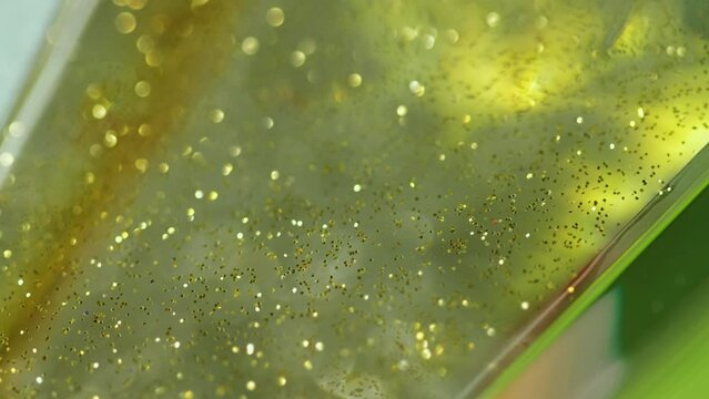 Glitter gel flow. Sparkling fluid. Defocused golden yellow green color shiny sequin particles transparent emulsion wave motion bokeh light texture abstract art background.