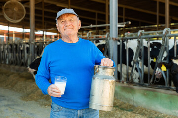 European old man farmer standing in cowshed with milk can and glass of milk in hands.
