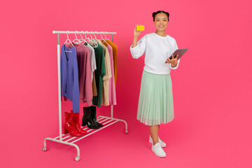 Asian woman holding credit card and digital tablet, standing near clothing rack