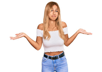 Beautiful blonde young woman wearing casual white tshirt clueless and confused expression with arms and hands raised. doubt concept.