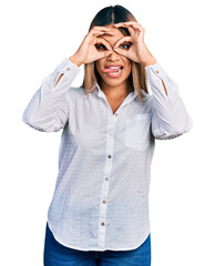 Young hispanic woman wearing casual white shirt doing ok gesture like binoculars sticking tongue out, eyes looking through fingers. crazy expression.