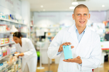 Male doctor in lab coat standing in drugstore with pharmaceutical package in hands. His co-worker setting out drugs in background.