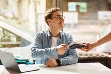 Cheerful young european man using credit card for payment machine, sit on desk with laptop