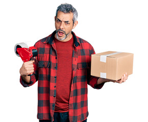 Middle age hispanic with grey hair holding packing tape and cardboard box in shock face, looking skeptical and sarcastic, surprised with open mouth