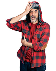 Middle age hispanic man with grey hair wearing fluffy earmuff hat surprised with hand on head for...