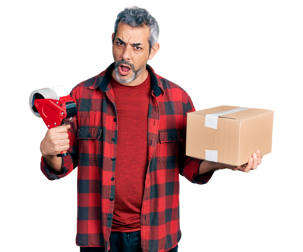 Middle age hispanic with grey hair holding packing tape and cardboard box in shock face, looking skeptical and sarcastic, surprised with open mouth