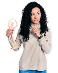 Young hispanic woman with curly hair holding small wooden manikin thinking concentrated about doubt with finger on chin and looking up wondering