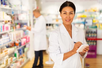 Zelfklevend Fotobehang Female Asian pharmacist in white gown standing in salesroom of chemist shop with pen and sheets of paper in hands and looking in camera. © JackF