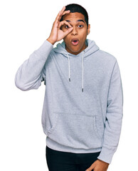 Young handsome hispanic man wearing casual sweatshirt doing ok gesture shocked with surprised face, eye looking through fingers. unbelieving expression.