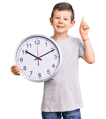 Cute blond kid holding big clock surprised with an idea or question pointing finger with happy...
