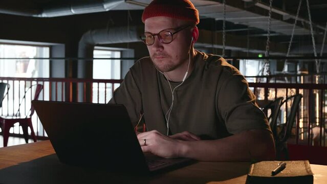 A millennial male IT programmer specialist is working at a laptop in white wired headphones, and is concentrating on the task at hand. A thoughtful freelancer in a dark room with a laptop. 4k footage