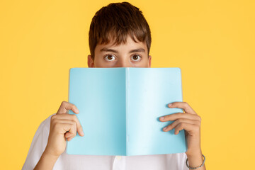 A boy with an intrigued expression carefully covers his face with a light blue book