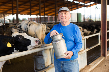 Old man farmer holding milk can in hands while standing in cowhouse.