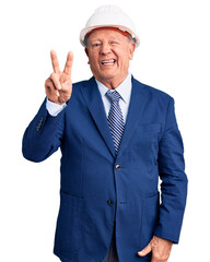 Senior handsome grey-haired man wearing suit and architect hardhat smiling with happy face winking at the camera doing victory sign. number two.
