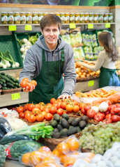 Positive young male seller holding tomatoes standing by counter in vegetable market