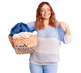 Young latin woman holding laundry basket pointing finger to one self smiling happy and proud