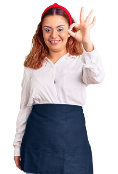 Young latin woman wearing waitress apron smiling positive doing ok sign with hand and fingers. successful expression.