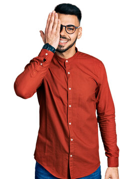 Young hispanic man with beard wearing business shirt and glasses covering one eye with hand, confident smile on face and surprise emotion.