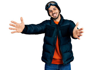 Young hispanic man with beard wearing snow wear and sky glasses looking at the camera smiling with open arms for hug. cheerful expression embracing happiness.